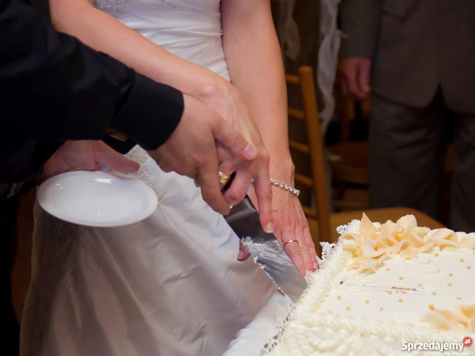 Para młoda kroi tort weselny zdobiony kremem i kwiatami, widoczne dłonie z obrączką i bransoletką oraz nóż do tortu.