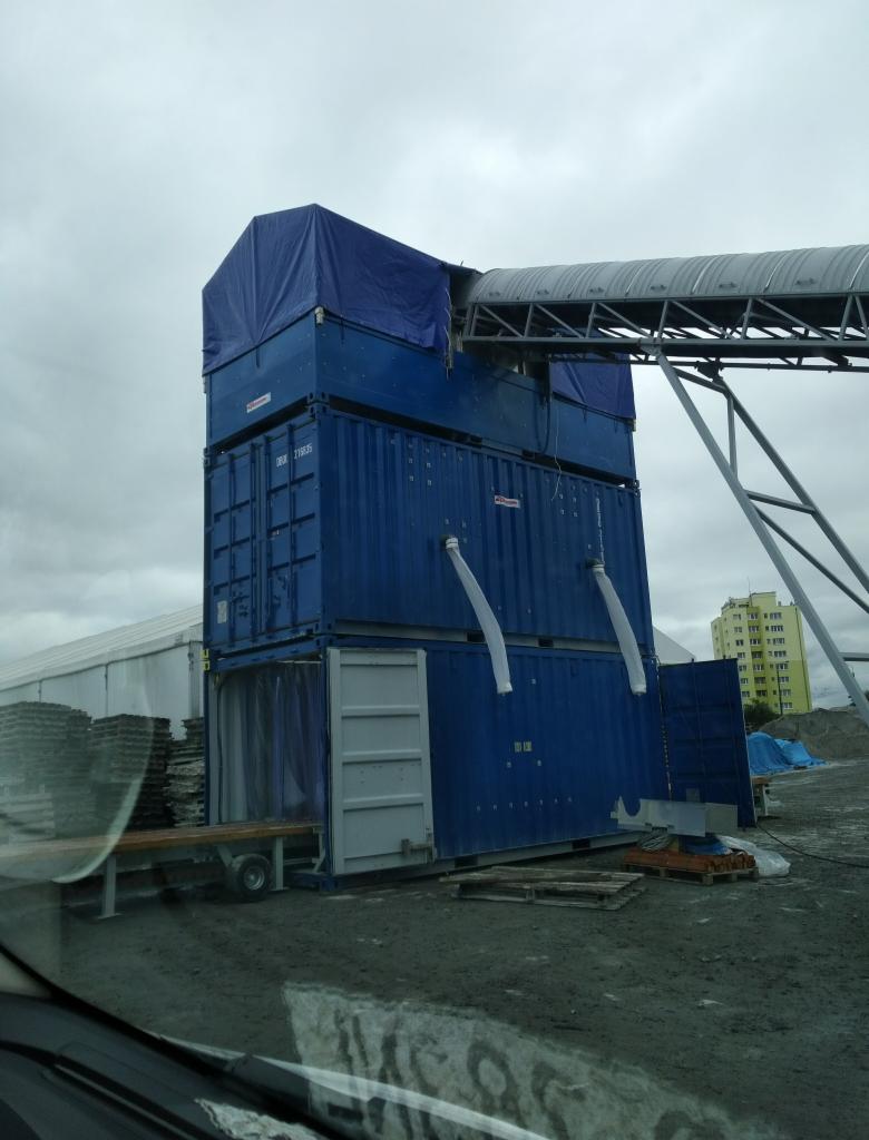 Trzy niebieskie kontenery ustawione jeden na drugim z otwartymi drzwiami na dole, połączone z metalową konstrukcją przypominającą tunel, całość przykryta plandeką, na szarym, błotnistym terenie.