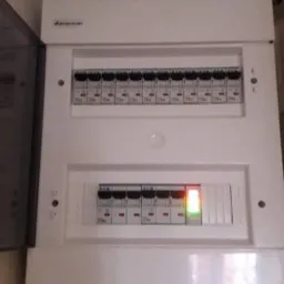 Biała skrzynka rozdzielcza z bezpiecznikami, jeden z nich sygnalizuje awarię czerwoną diodą, widoczna otwarta obudowa i fragment rury na tle ściany.