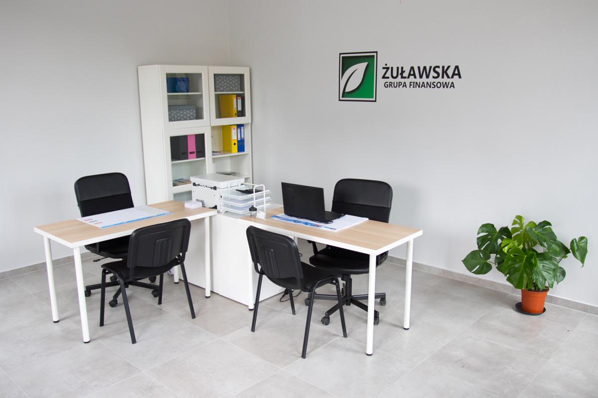 Jasne biuro z dwoma biurkami, krzesłami, szafką na dokumenty, laptopem, dokumentami i rośliną doniczkową, na ścianie logo firmy Żuławska Grupa Finansowa.