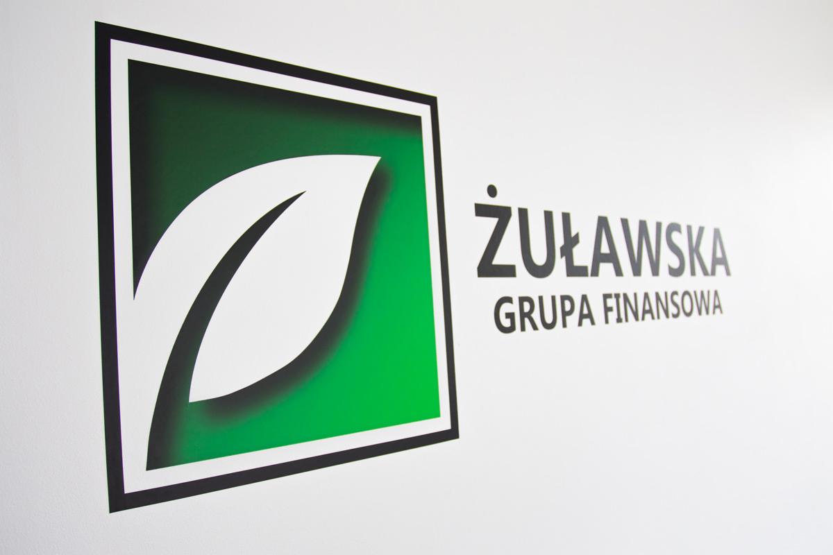 Logo Żuławska Grupa Finansowa z zielonym liściem w kwadracie, umieszczone na białej ścianie.