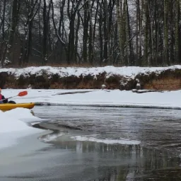 Kajakarz w pomarańczowej czapce płynie żółtym kajakiem po rzece częściowo zamarzniętej, z brzegami pokrytymi śniegiem i lasem w tle.