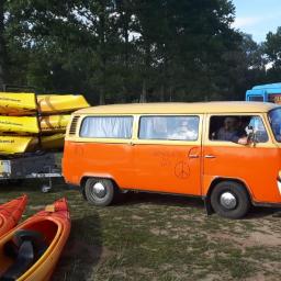 FHU Kolor Magdalena Wolak - Pomarańczowy bus z napisem 'Noce Bolata Tu Była' i symbolem pacyfki, ciągnący przyczepę z żółtymi kajakami, na trawie leżą czerwone kajaki, w tle niebieski bus z reklamą.