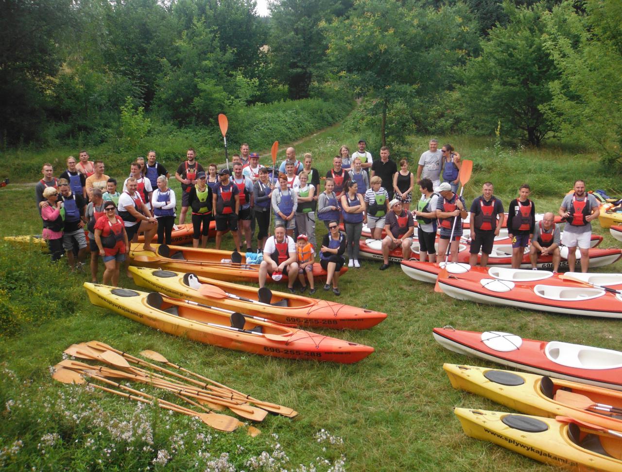 Grupa osób w kamizelkach asekuracyjnych pozuje na trawie obok kajaków i wioseł przed wycieczką kajakową, na kajakach widoczne numery kontaktowe.