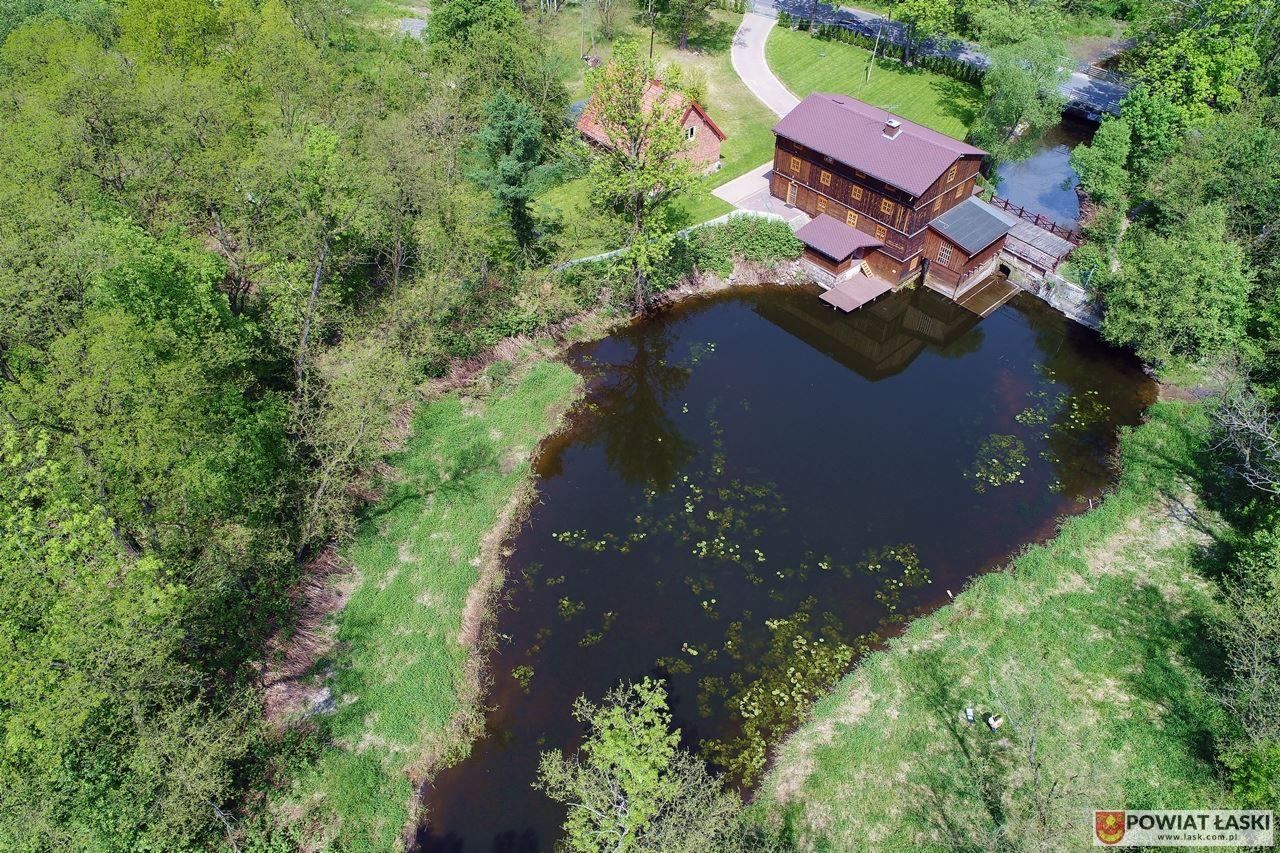 Widok z drona na stary młyn wodny z czerwonym dachem, staw z liliami wodnymi i otaczającą zieleń, tworzący malowniczy krajobraz.
