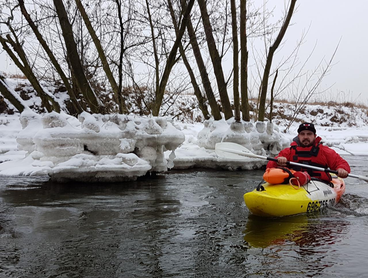 Mężczyzna w czerwonej kurtce płynie kajakiem po rzece w zimowej scenerii, z brzegami pokrytymi śniegiem i lodem.
