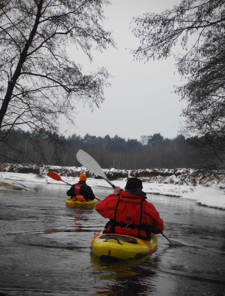 Dwaj kajakarze płyną żółtymi kajakami po rzece w zimowej scenerii, brzeg pokryty śniegiem, widoczne gołe drzewa i las w tle.
