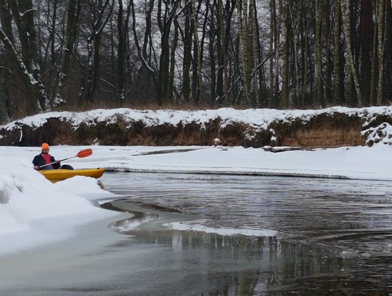 Kajakarz w pomarańczowej czapce płynie żółtym kajakiem po rzece częściowo zamarzniętej, z brzegami pokrytymi śniegiem i lasem w tle.
