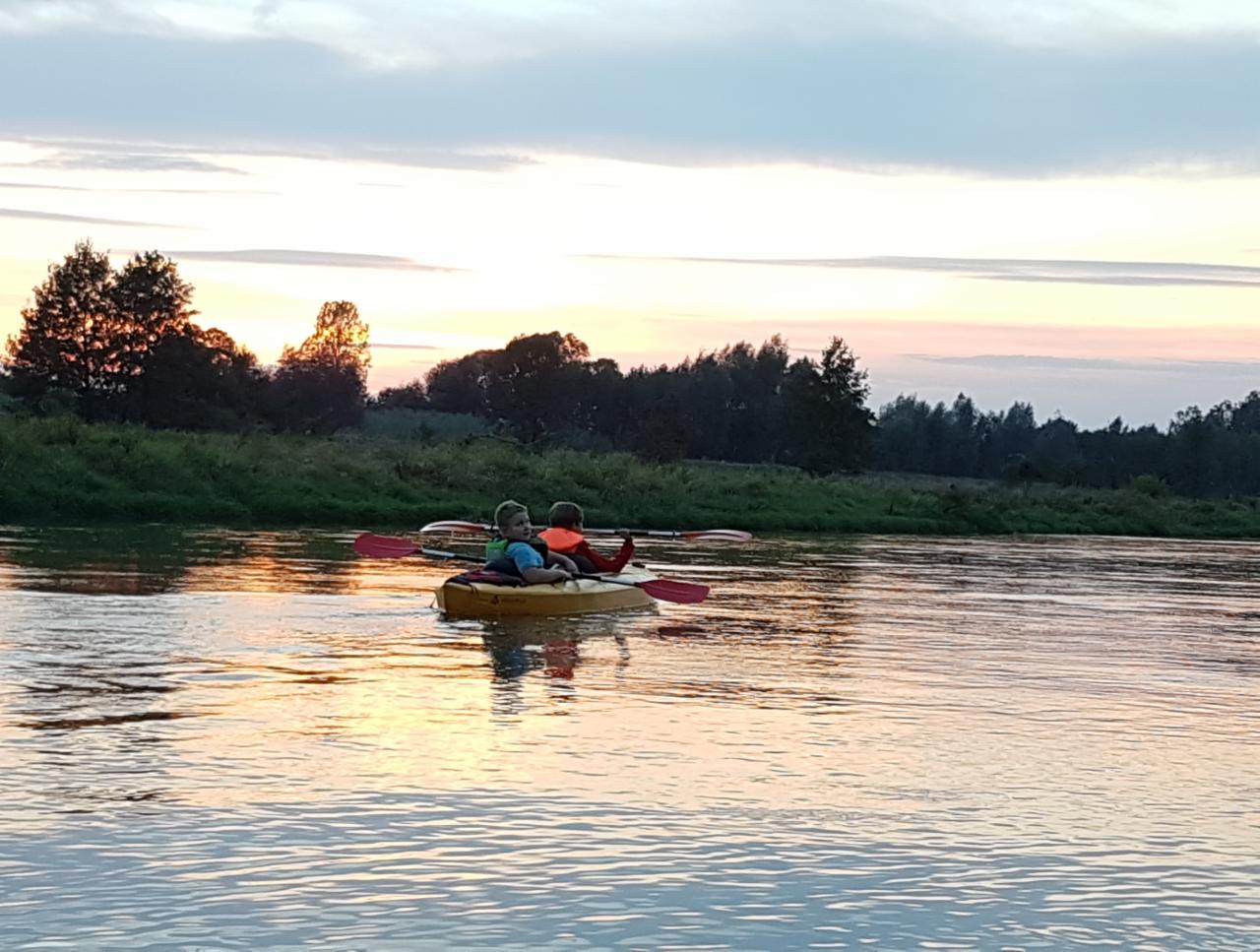 Dwoje dzieci w kajaku na rzece o zmierzchu, odbicie zachodzącego słońca w wodzie, drzewa na brzegu w tle.