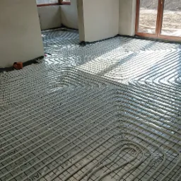 Rozłożone rury ogrzewania podłogowego w surowym wnętrzu, widoczne okna i przygotowane podłoże z folią izolacyjną.