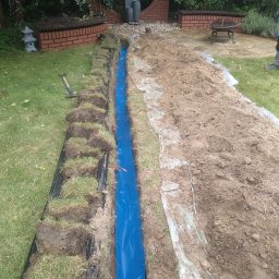 WD ELEKTRO - Wykopany rów z niebieską folią w ogrodzie, trawa z ziemią obok, w tle elementy dekoracyjne ogrodu i grządki.