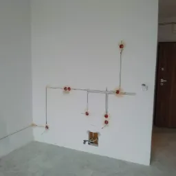 Wnętrze w stanie deweloperskim z widoczną instalacją elektryczną: kable poprowadzone po ścianach, puszki elektryczne zamontowane w otworach, brak osprzętu, surowy beton na podłodze, drewniane drzwi...