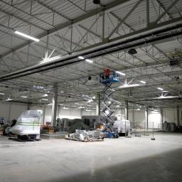 WD ELEKTRO - Pracownik w pomarańczowej kamizelce na podnośniku nożycowym montuje instalacje pod sufitem hali przemysłowej z widocznymi lampami i przewodami.