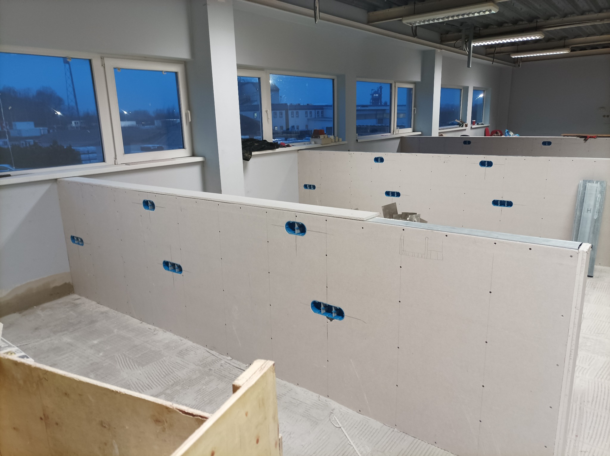 Wnętrze biurowe w trakcie budowy, widoczne ściany działowe z płyt kartonowo-gipsowych z przygotowanymi otworami na gniazdka elektryczne, surowy beton na podłodze, okna z widokiem na zewnątrz.