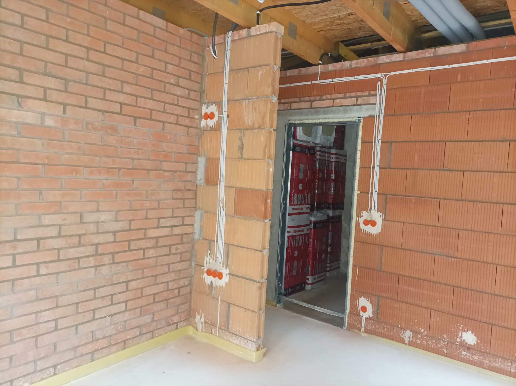 Surowy mur z cegły z widocznymi, świeżo poprowadzonymi przewodami elektrycznymi i pomarańczowymi puszkami instalacyjnymi, widok na nieotynkowane wnętrze z zapakowanymi materiałami budowlanymi w tle.