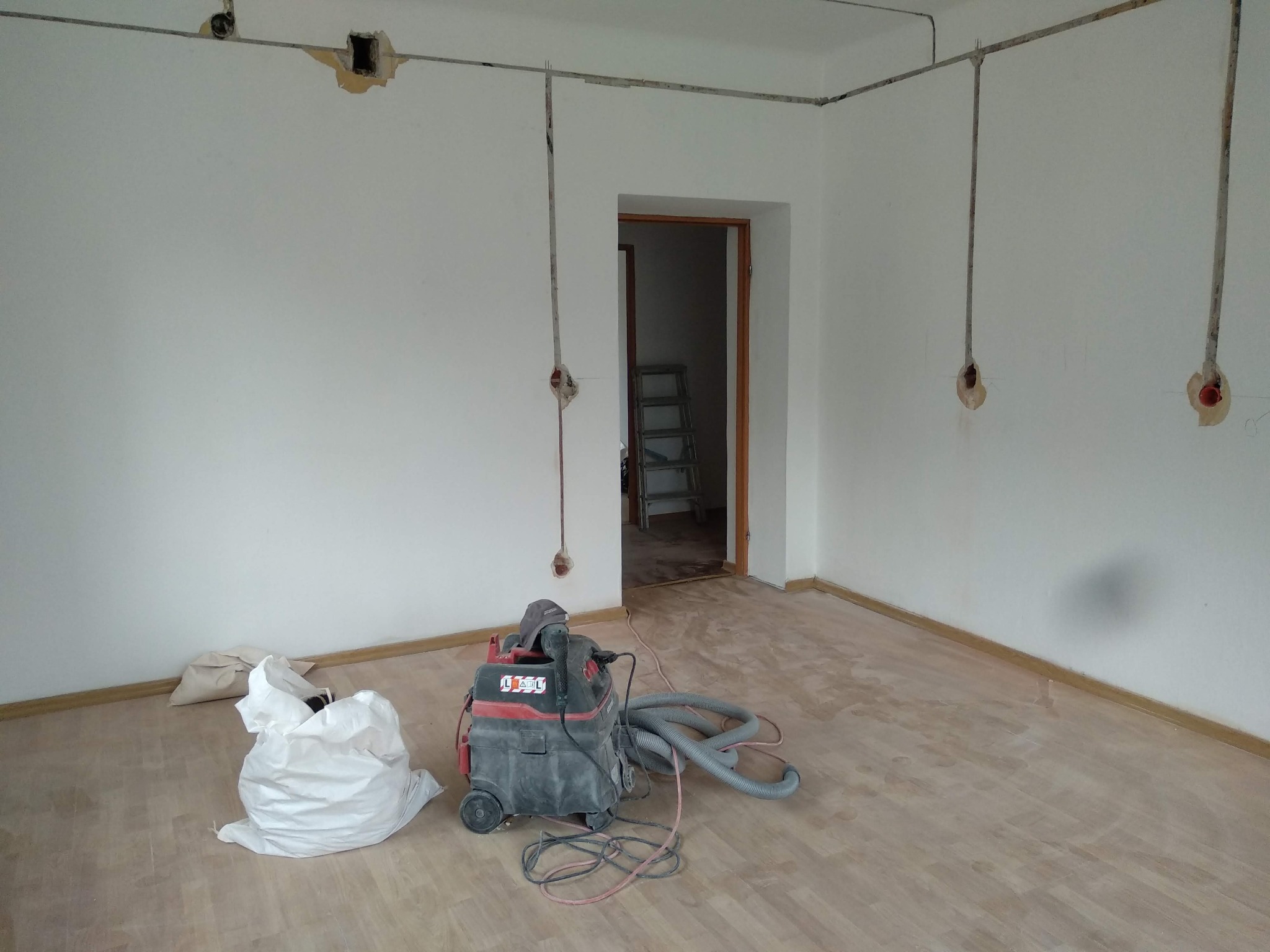 Wnętrze w trakcie remontu: widoczne przewody elektryczne wystające ze ścian, otwory pod gniazdka, odkurzacz przemysłowy na podłodze i worki z odpadami.