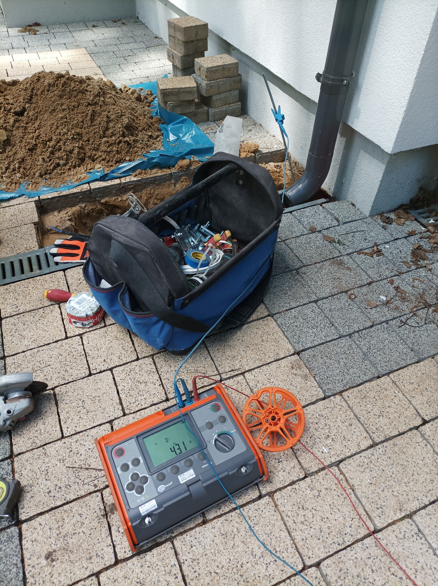 Miernik rezystancji uziemień marki Sonel MRU-1 na kostce brukowej, obok torba narzędziowa, szlifierka kątowa i wykop z piaskiem zabezpieczony folią podczas pomiaru uziomu przy rurze spustowej.