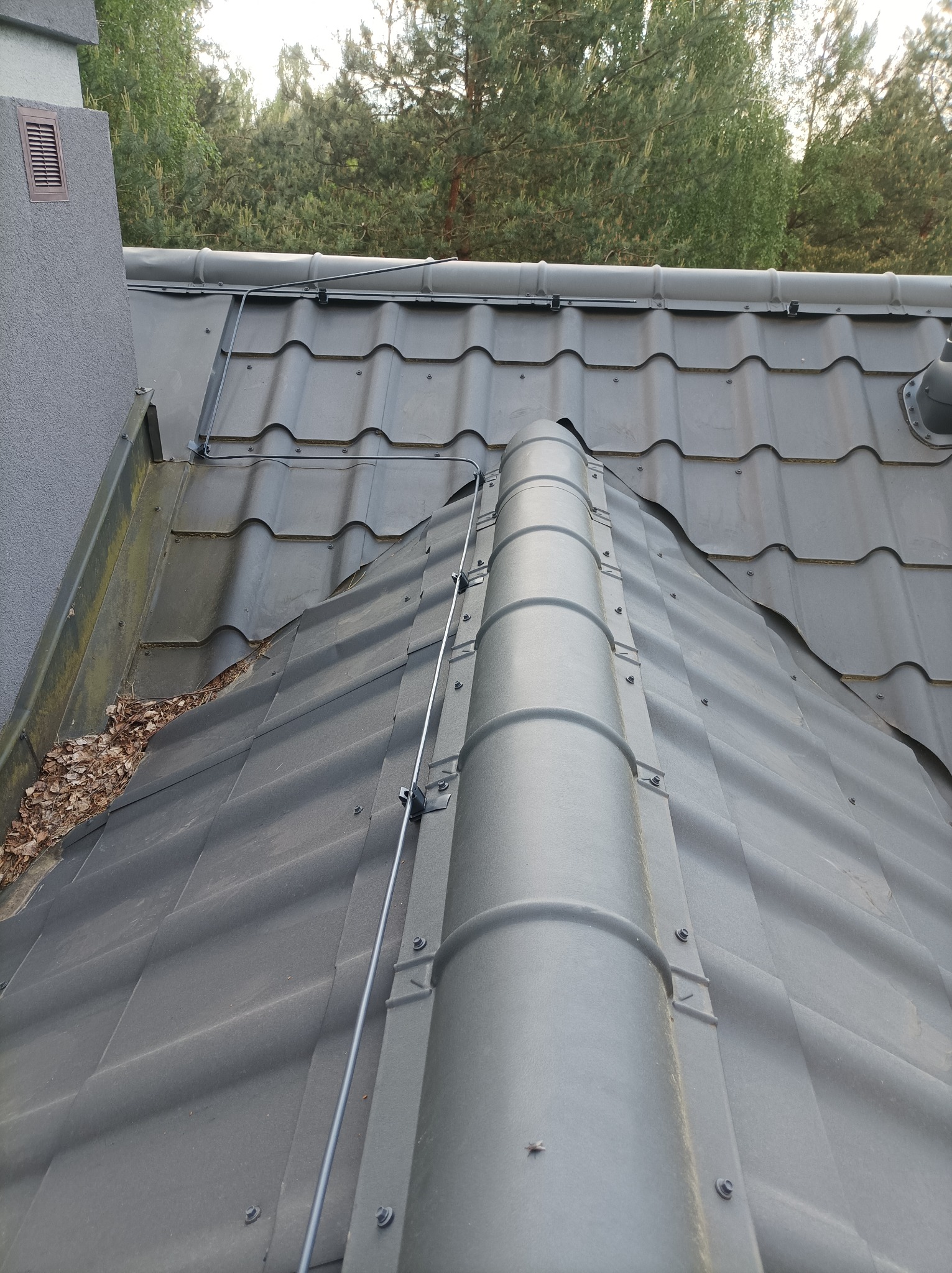 Szara blachodachówka z zamontowaną instalacją odgromową, widoczne mocowania i przewody, na dachu liście w rynnie