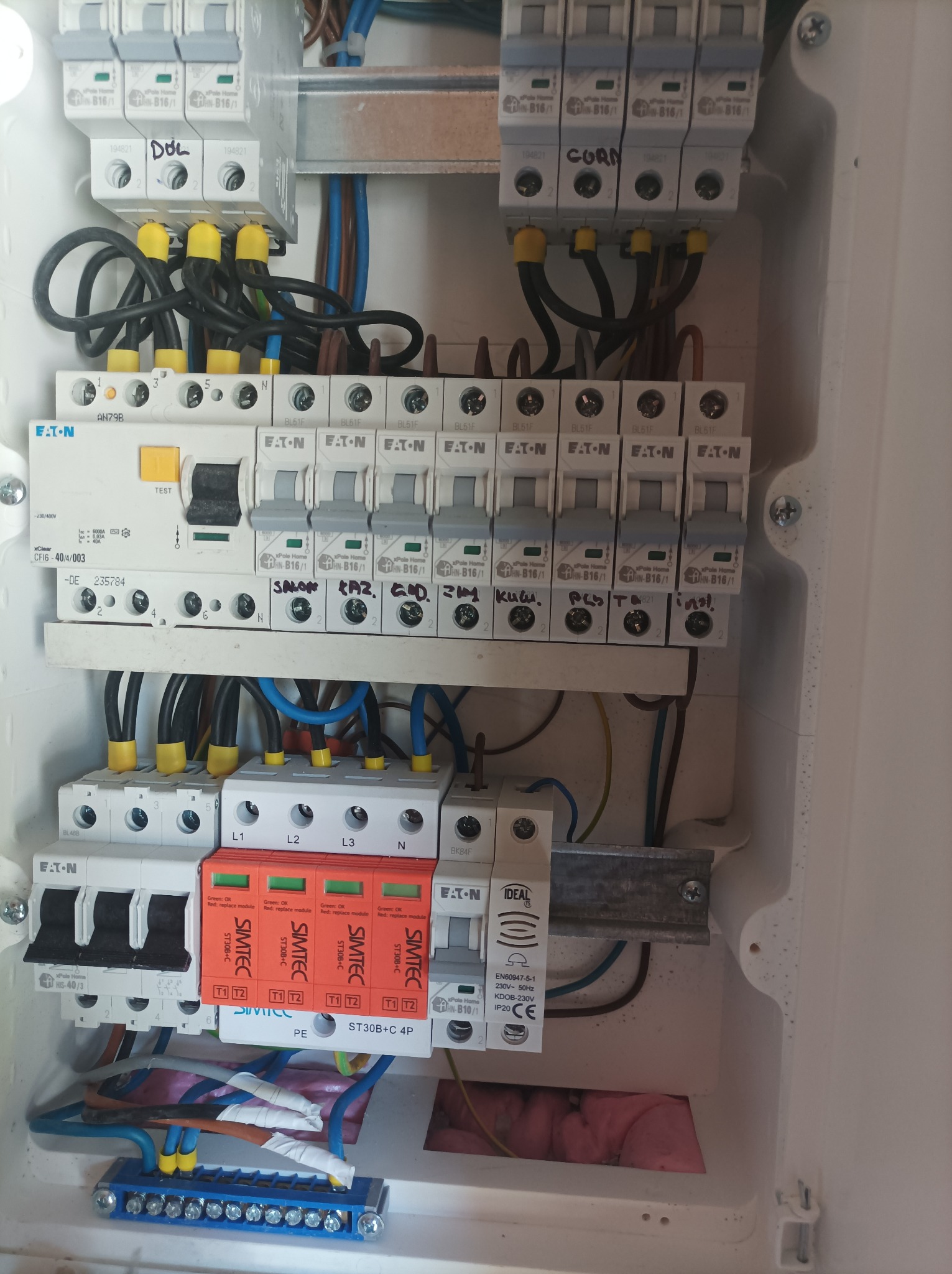 Szczegółowe ujęcie wnętrza skrzynki elektrycznej z zamontowanymi bezpiecznikami, ogranicznikami przepięć Simtec i listwą zaciskową, widoczne przewody w różnych kolorach z żółtymi osłonkami.