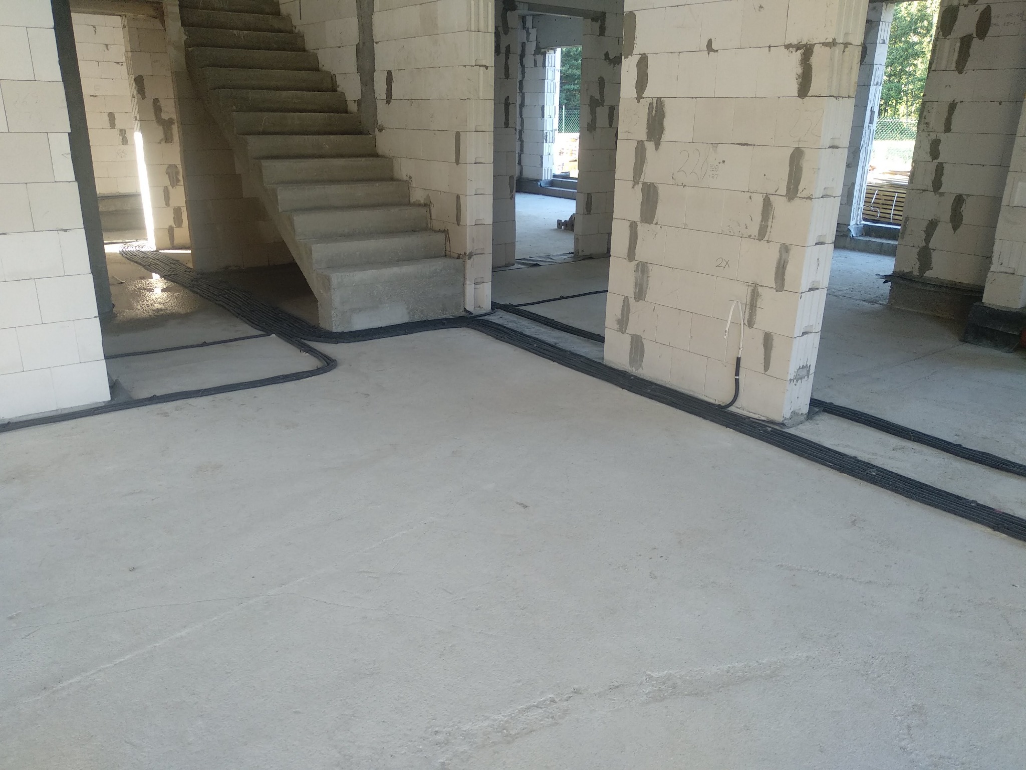 Surowy stan budynku w trakcie budowy z widocznymi kablami elektrycznymi ułożonymi na podłodze, prowadzącymi wzdłuż ścian i pod schodami, ściany z białych bloczków.