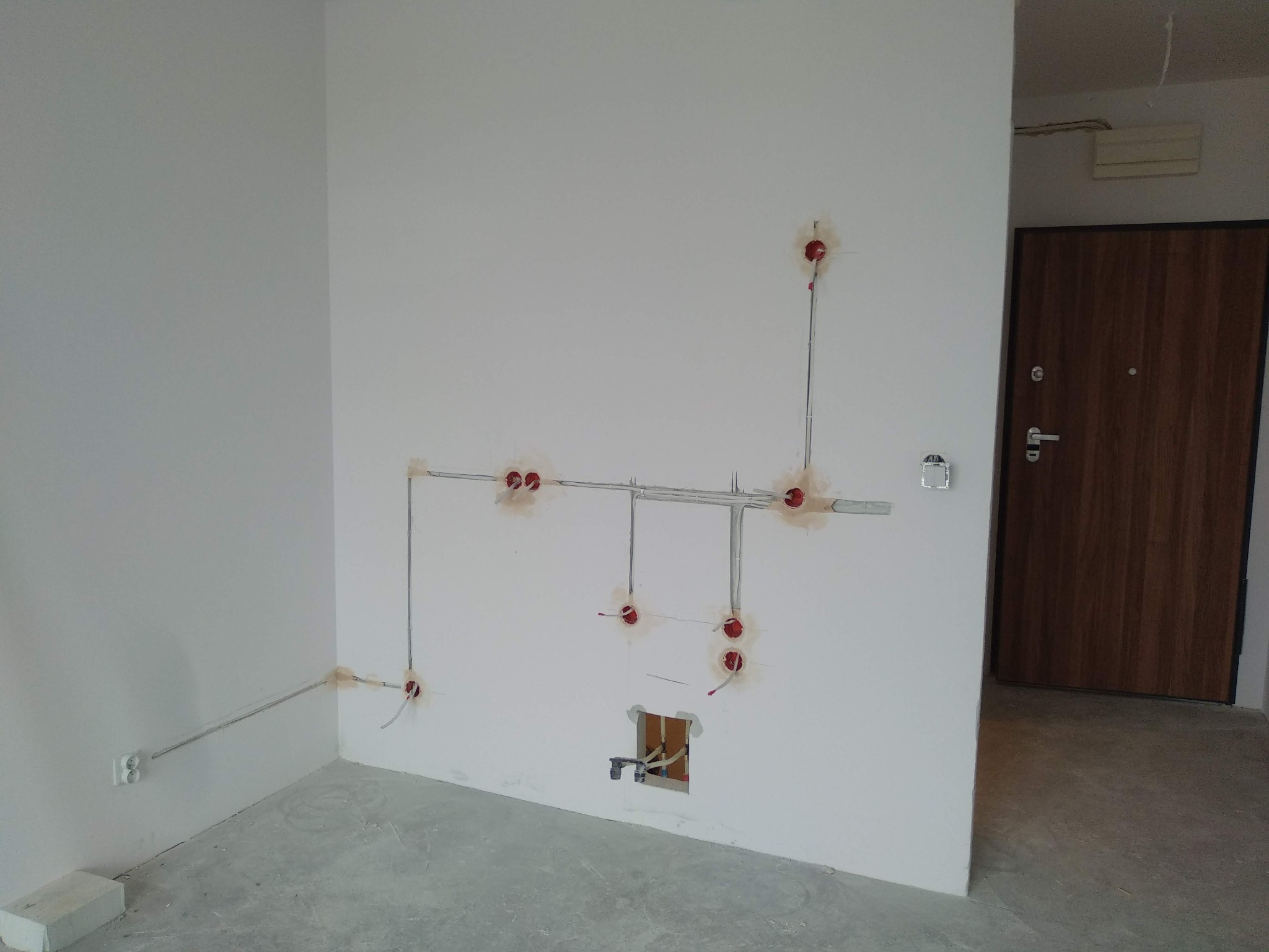 Wnętrze w stanie deweloperskim z widoczną instalacją elektryczną: kable poprowadzone po ścianach, puszki elektryczne zamontowane w otworach, brak osprzętu, surowy beton na podłodze, drewniane drzwi...