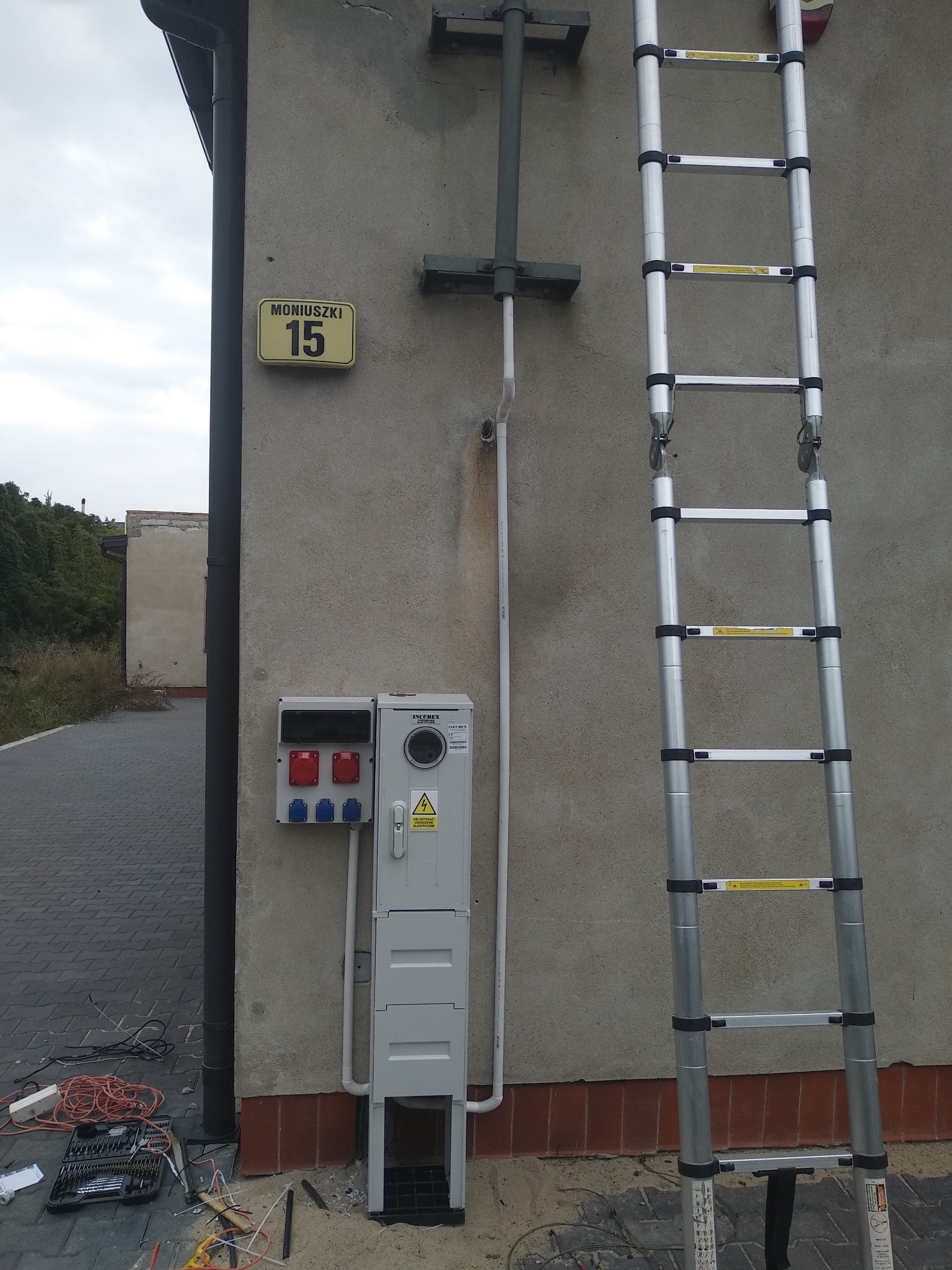 Szara skrzynka elektryczna z czerwonymi i niebieskimi gniazdami oraz tabliczką ostrzegawczą, zamontowana na elewacji budynku obok numeru domu 'Moniuszki 15', z rozłożoną aluminiową drabiną...