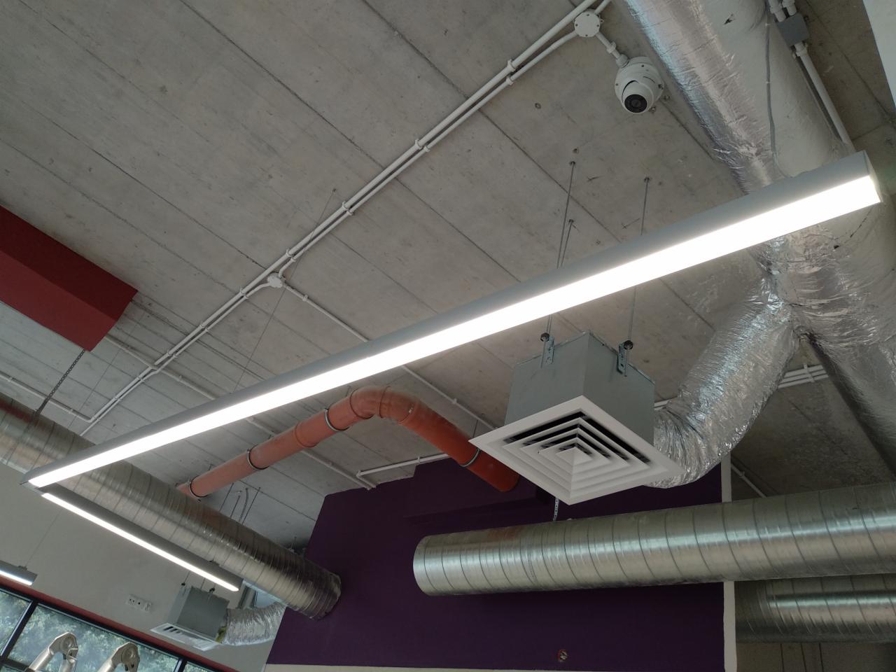 Widok sufitu w hali przemysłowej z widocznymi instalacjami: rurami wentylacyjnymi, kanałami wentylacyjnymi z kratkami, oświetleniem liniowym LED, przewodami w białych listwach oraz kamerą przemysłową.
