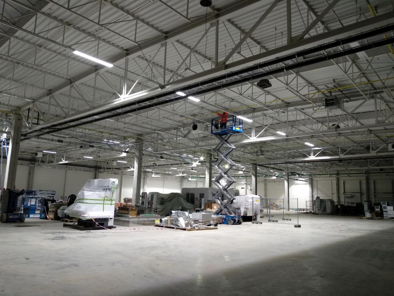 Pracownik w pomarańczowej kamizelce na podnośniku nożycowym montuje instalacje pod sufitem hali przemysłowej z widocznymi lampami i przewodami.