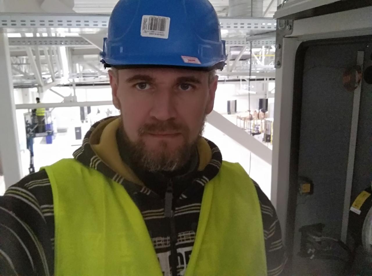 Elektryk w niebieskim kasku z naklejką i żółtej kamizelce odblaskowej pozuje na tle surowego wnętrza komercyjnego budynku z widoczną instalacją elektryczną i skrzynką rozdzielczą