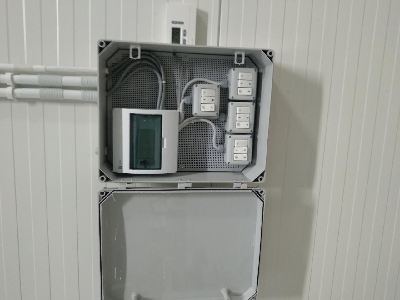 Otwarta, szara skrzynka elektryczna z zainstalowanym rozdzielnicą, czterema włącznikami i widocznymi przewodami na tle białej ściany z panelami.