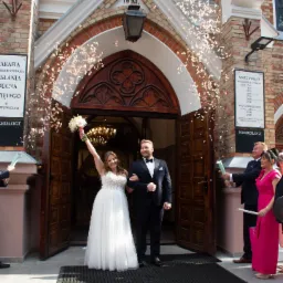 Para Młoda wychodzi z Kościoła w deszczu konfetti. Panna Młoda z uniesionym bukietem, uśmiechnięta. Goście weselni z tubami konfetti. Budynek kościoła z cegły.