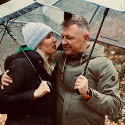Magdalena Pawlak - Uśmiechnięta para pod parasolami w deszczowy dzień. Kobieta w czapce i kurtce, mężczyzna w zielonej kurtce. Jesienne liście w tle, bliski kontakt wzrokowy.