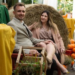 Magdalena Pawlak - Uśmiechnięta para w jesiennej scenerii: mężczyzna w beżowym garniturze, kobieta w sukience i płaszczu, siedzą na tle siana i dyń. Romantyczna sesja plenerowa.