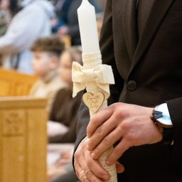 Magdalena Pawlak - Dłoń mężczyzny w garniturze trzyma świecę do chrztu z ozdobną kokardą i drewnianym sercem z napisem na tle kościelnych ławek i rozmytych postaci.