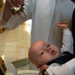 Magdalena Pawlak - Chrzest dziecka: woda z naczynia polewana na główkę niemowlęcia trzymanego przez mężczyznę w białej szacie. Ujęcie z bliska, widoczna twarz dziecka i dłoń z naczyniem.