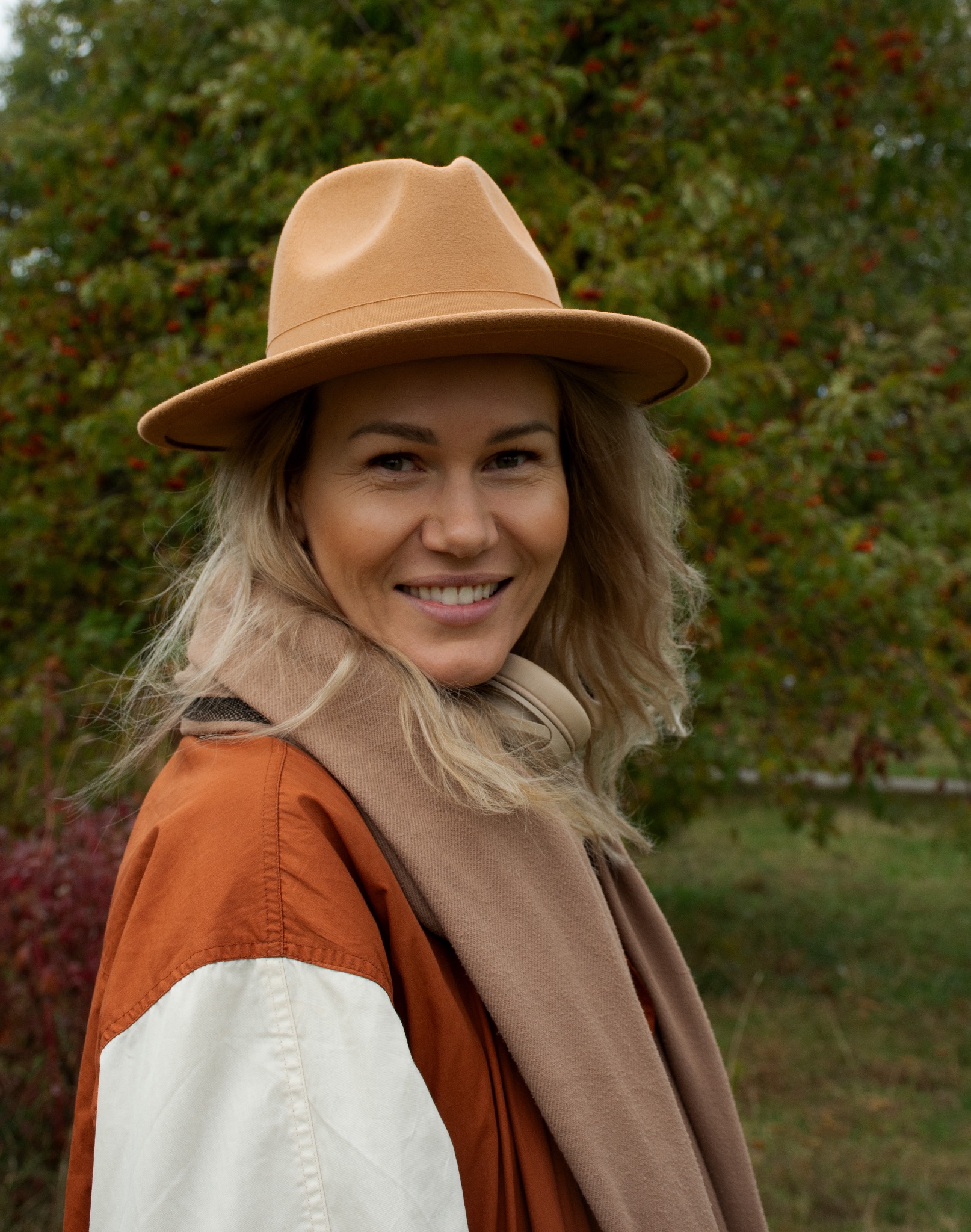 Uśmiechnięta blondynka w kapeluszu i szaliku na tle zieleni. Portret w plenerze, ciepłe kolory, naturalne światło. Widoczne słuchawki na szyi.