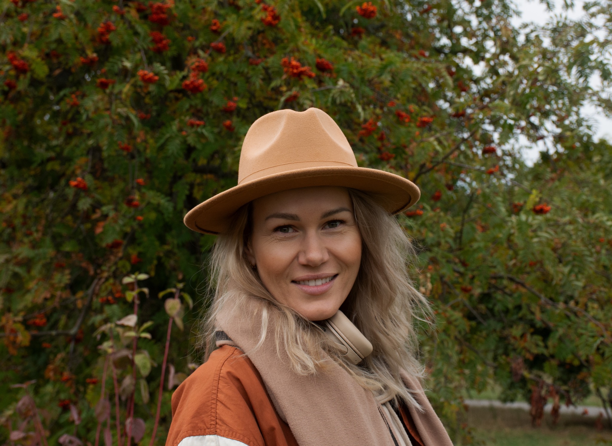 Portret uśmiechniętej blondynki w kapeluszu i szaliku na tle drzewa z jarzębiną. Jesienna stylizacja, naturalne światło, ciepłe barwy.