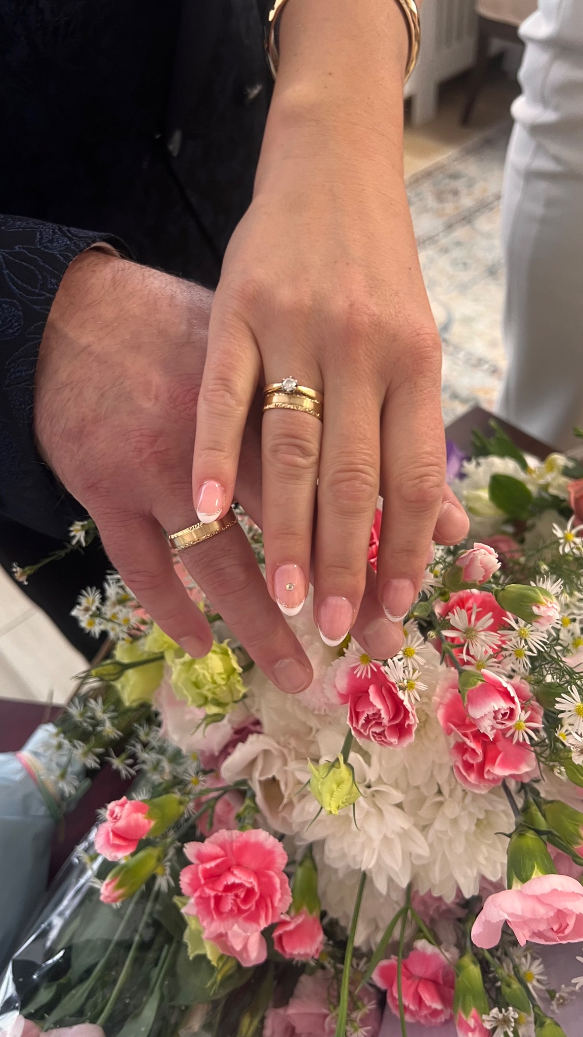 Zbliżenie dłoni pary młodej z obrączkami na tle bukietu kwiatów. Kobieta ma manicure francuski z delikatnym zdobieniem. Ujęcie z góry, ciepłe światło.