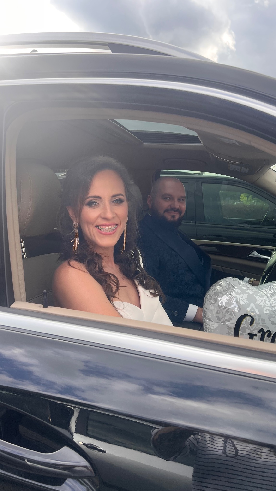 Uśmiechnięta kobieta w białej sukience i z aparatem ortodontycznym siedzi w samochodzie obok mężczyzny w garniturze, trzymając balon z napisem 'Gratulacje'.