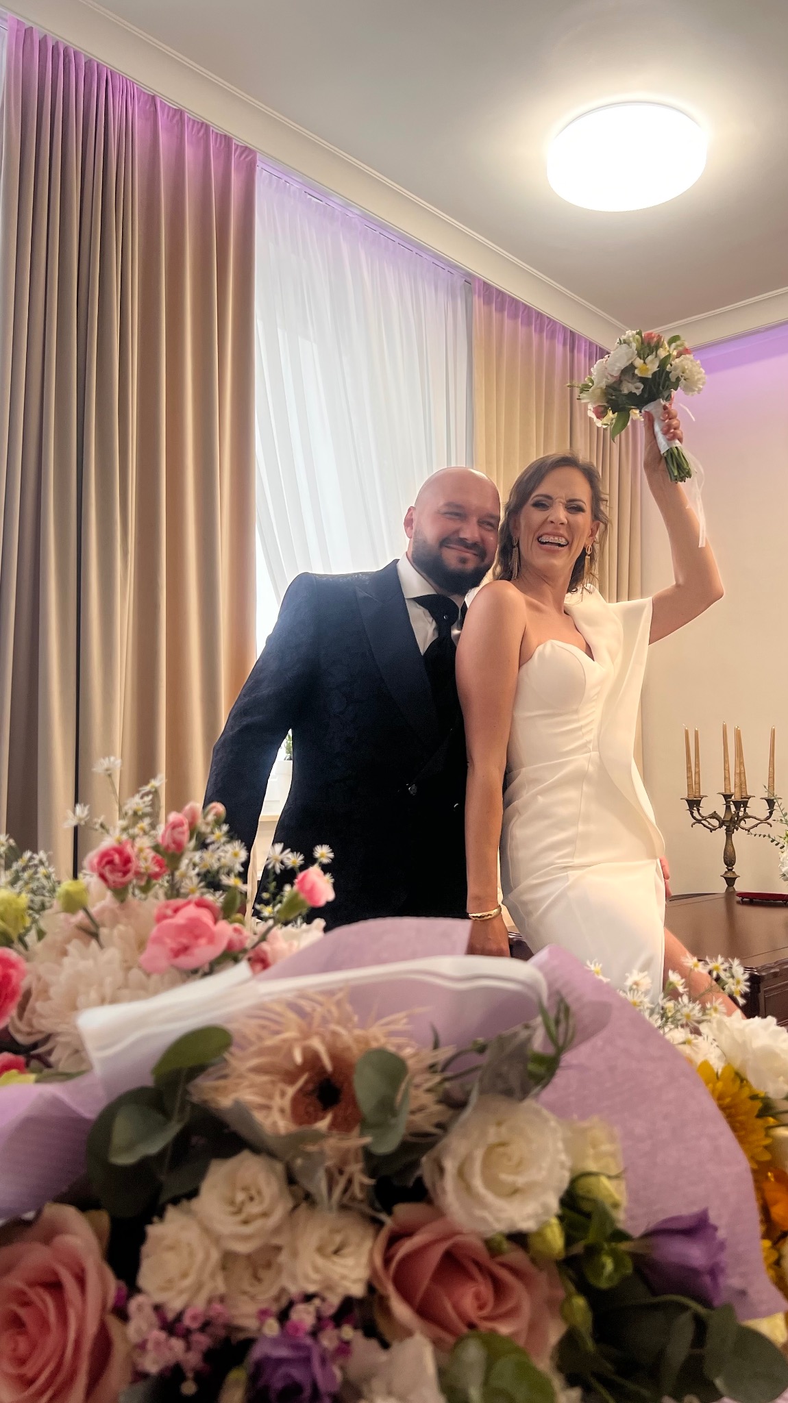 Uśmiechnięta para młoda w eleganckich strojach pozuje z bukietem kwiatów na tle jasnych zasłon i dekoracji kwiatowych. Radosna atmosfera.