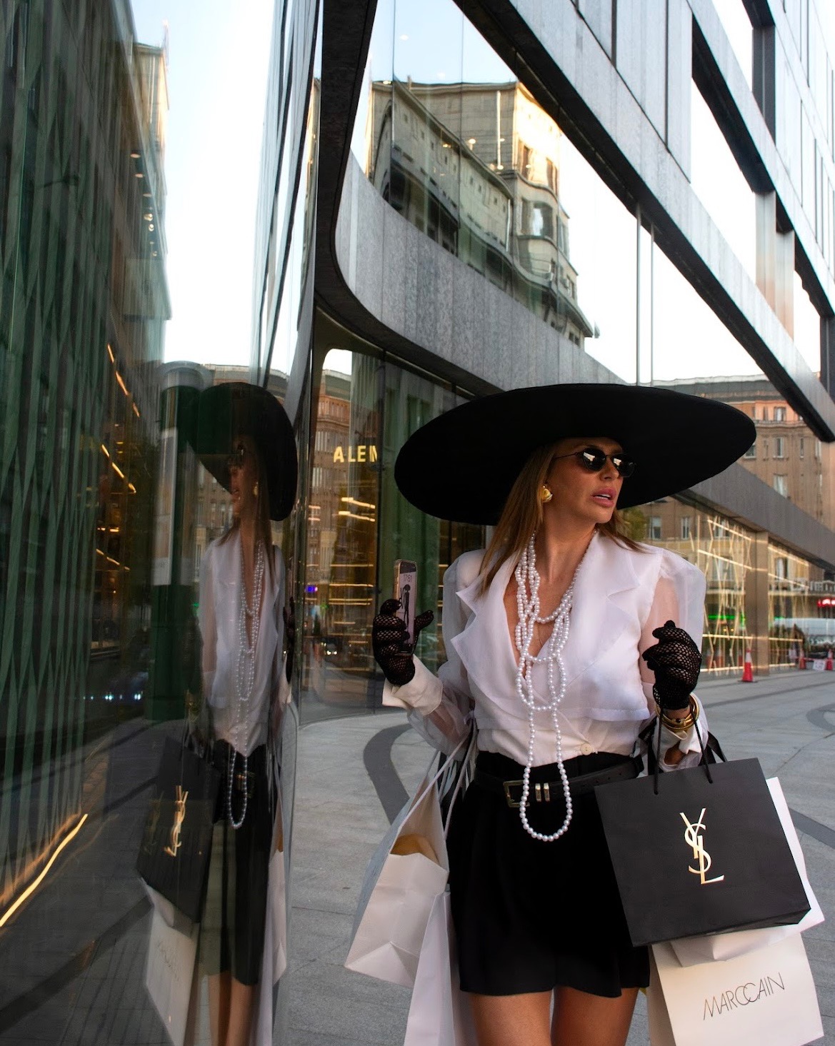 Elegancka kobieta w kapeluszu i okularach przeciwsłonecznych, z torbami na zakupy, pozuje na tle nowoczesnej architektury miejskiej. Widoczne odbicie w szklanej fasadzie.