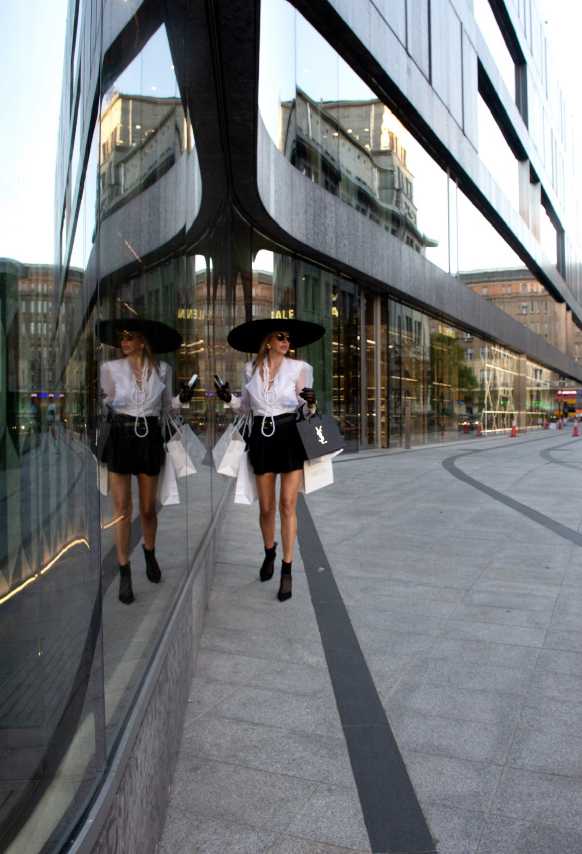 Elegancka kobieta w kapeluszu i okularach przeciwsłonecznych, niosąca torby na zakupy, odbija się w szklanej fasadzie nowoczesnego budynku.