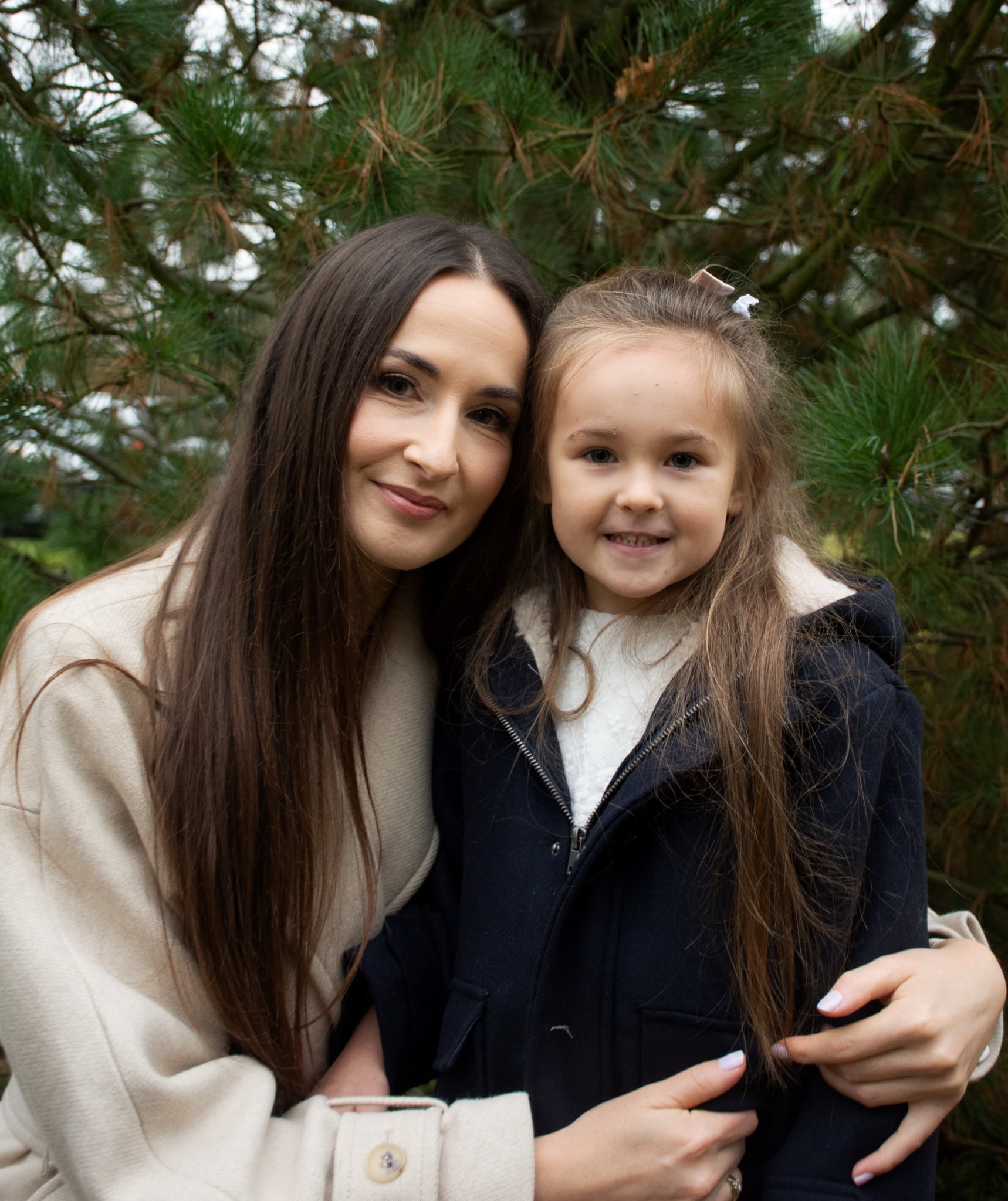 Portret uśmiechniętej matki z córką na tle zielonych gałęzi drzewa iglastego. Kobiety w zimowych płaszczach, bliski kadr, naturalne światło.