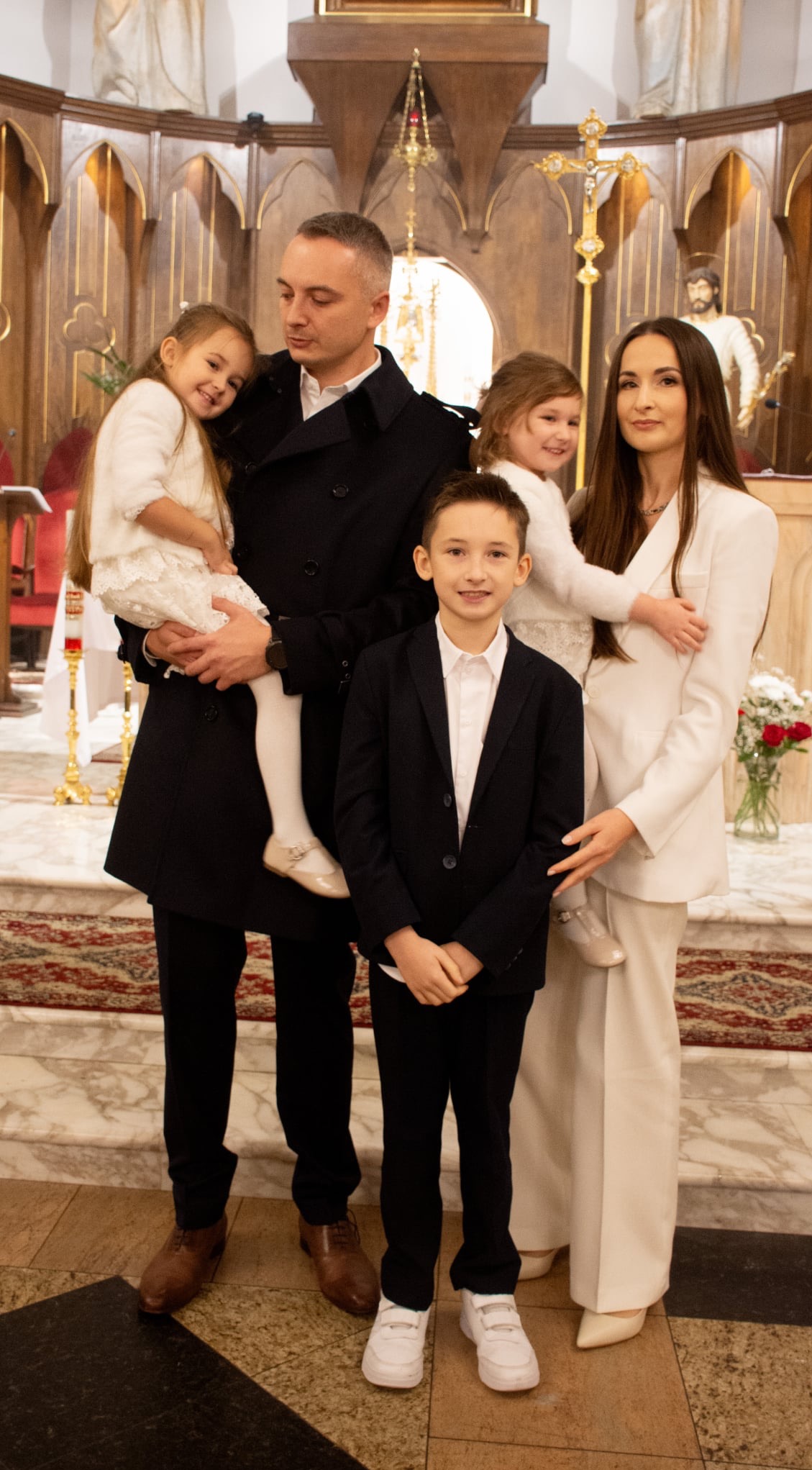 Uroczysta fotografia rodzinna w kościele: rodzice z trójką dzieci, eleganckie stroje, marmurowa posadzka, dekoracje sakralne w tle. Pamiątka chrztu lub innego ważnego wydarzenia.