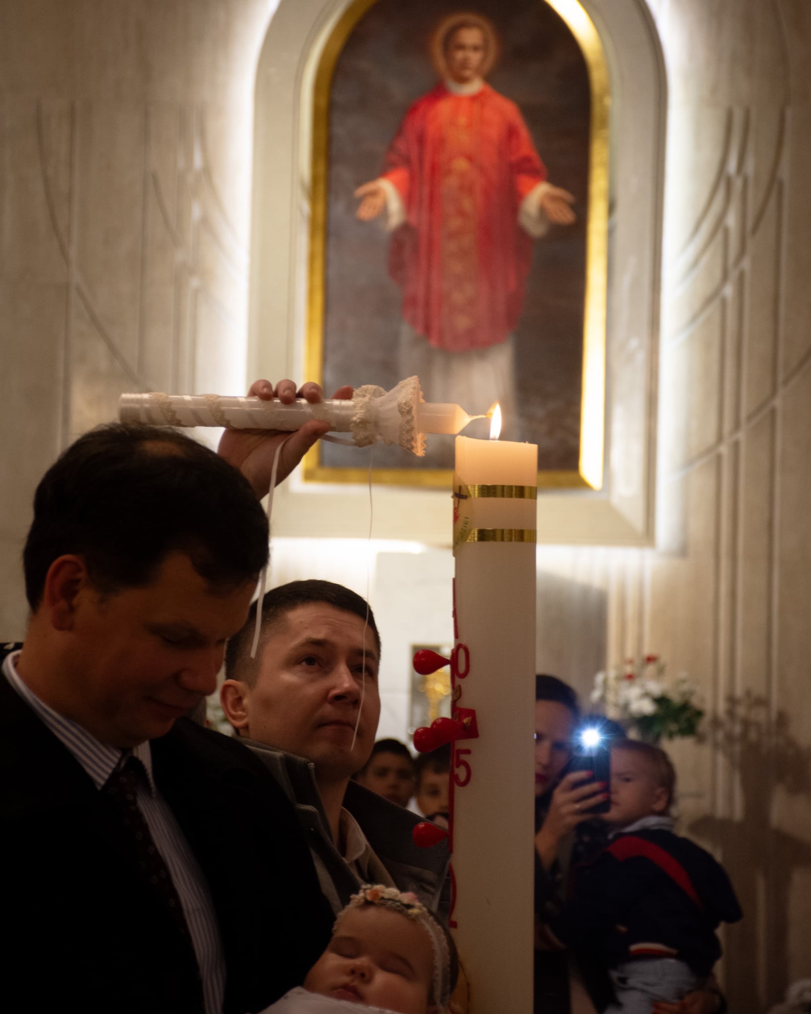 Chrzest dziecka w kościele: ojciec chrzestny zapala świecę, w tle obraz religijny i goście. Ujęcie z bliska, ciepłe światło świec.