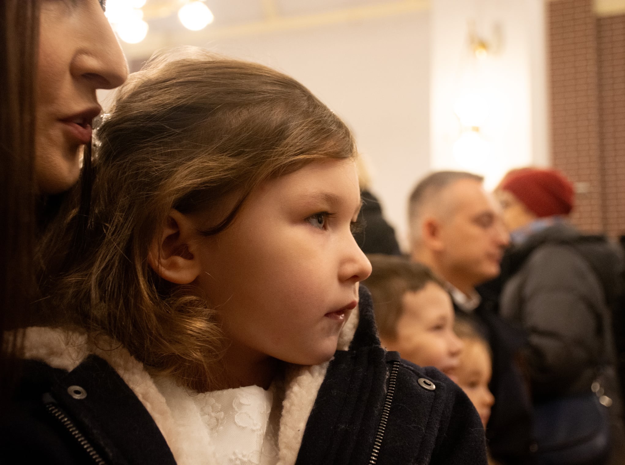Profil dziewczynki w białej sukience i granatowej kurtce na tle innych osób. Ujęcie z bliska, ciepłe oświetlenie, skupienie na detalu twarzy dziecka.
