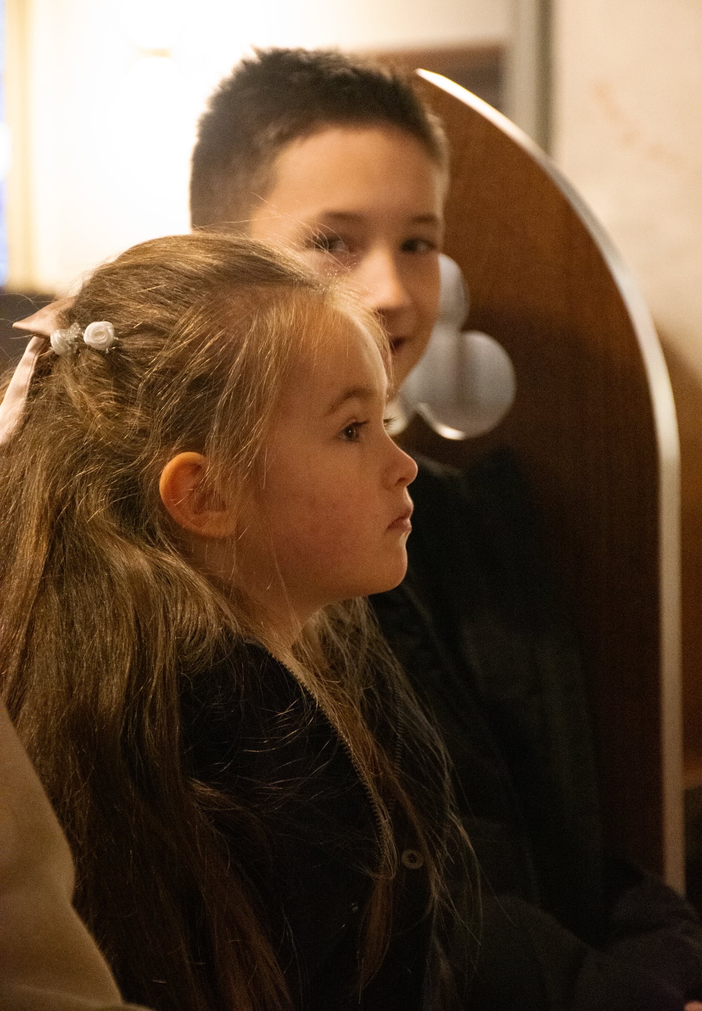 Portret profilowy dziewczynki z długimi włosami ozdobionymi kwiatami, w tle widoczny chłopiec, oboje w kościele. Ciepłe, stonowane oświetlenie.