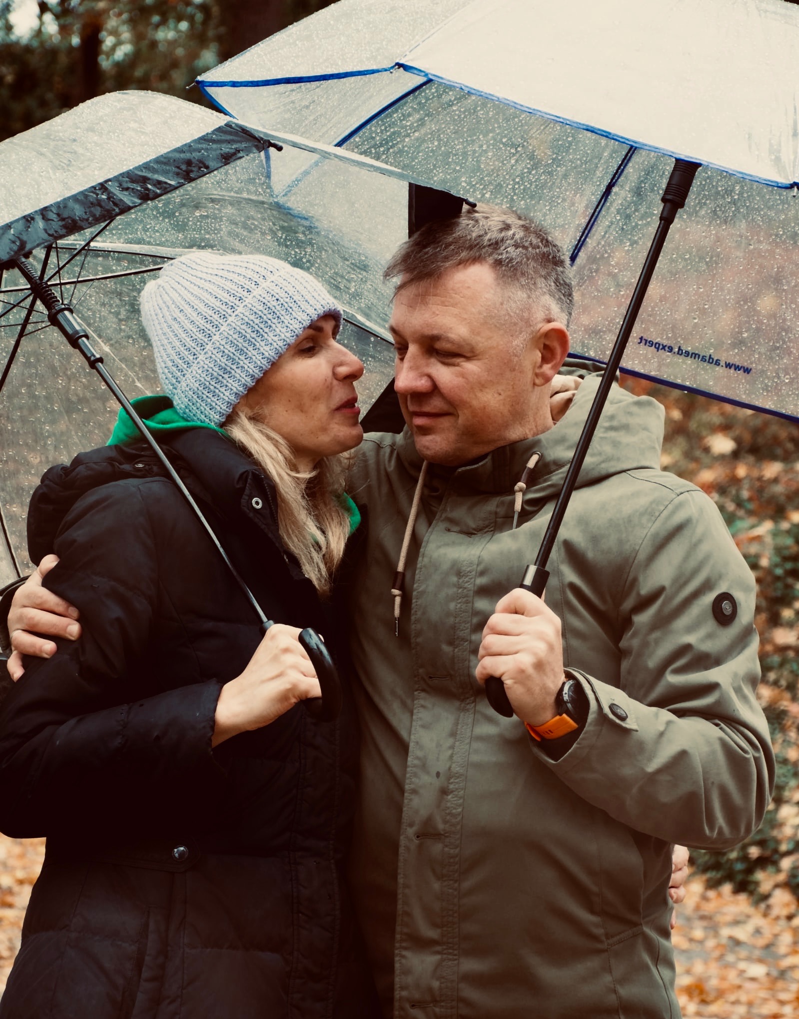 Uśmiechnięta para pod parasolami w deszczowy dzień. Kobieta w czapce i kurtce, mężczyzna w zielonej kurtce. Jesienne liście w tle, bliski kontakt wzrokowy.