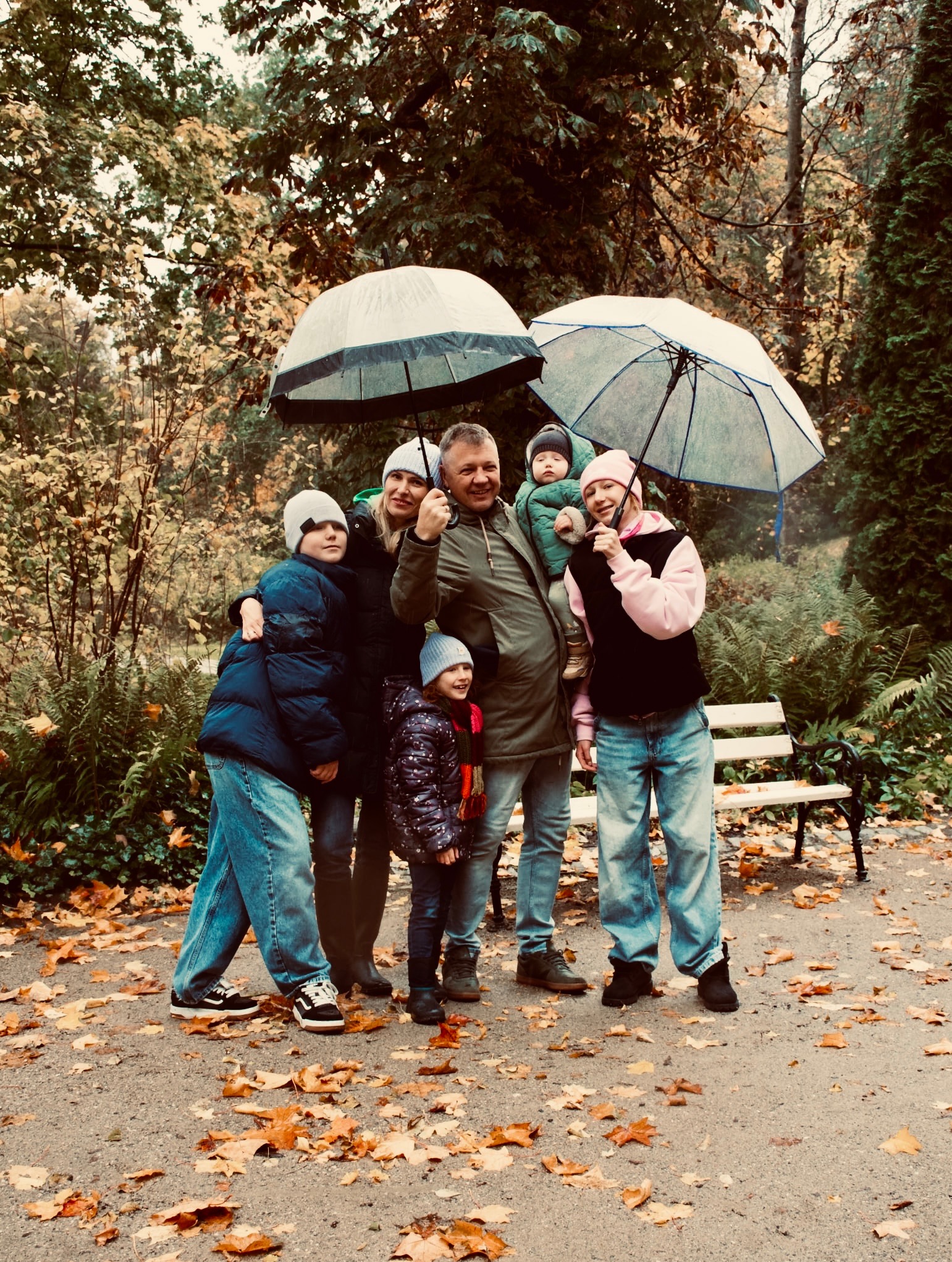 Jesienna sesja rodzinna w parku: uśmiechnięta grupa pod parasolami, kolorowe liście na ziemi, ławka w tle. Naturalne światło, swobodna atmosfera.