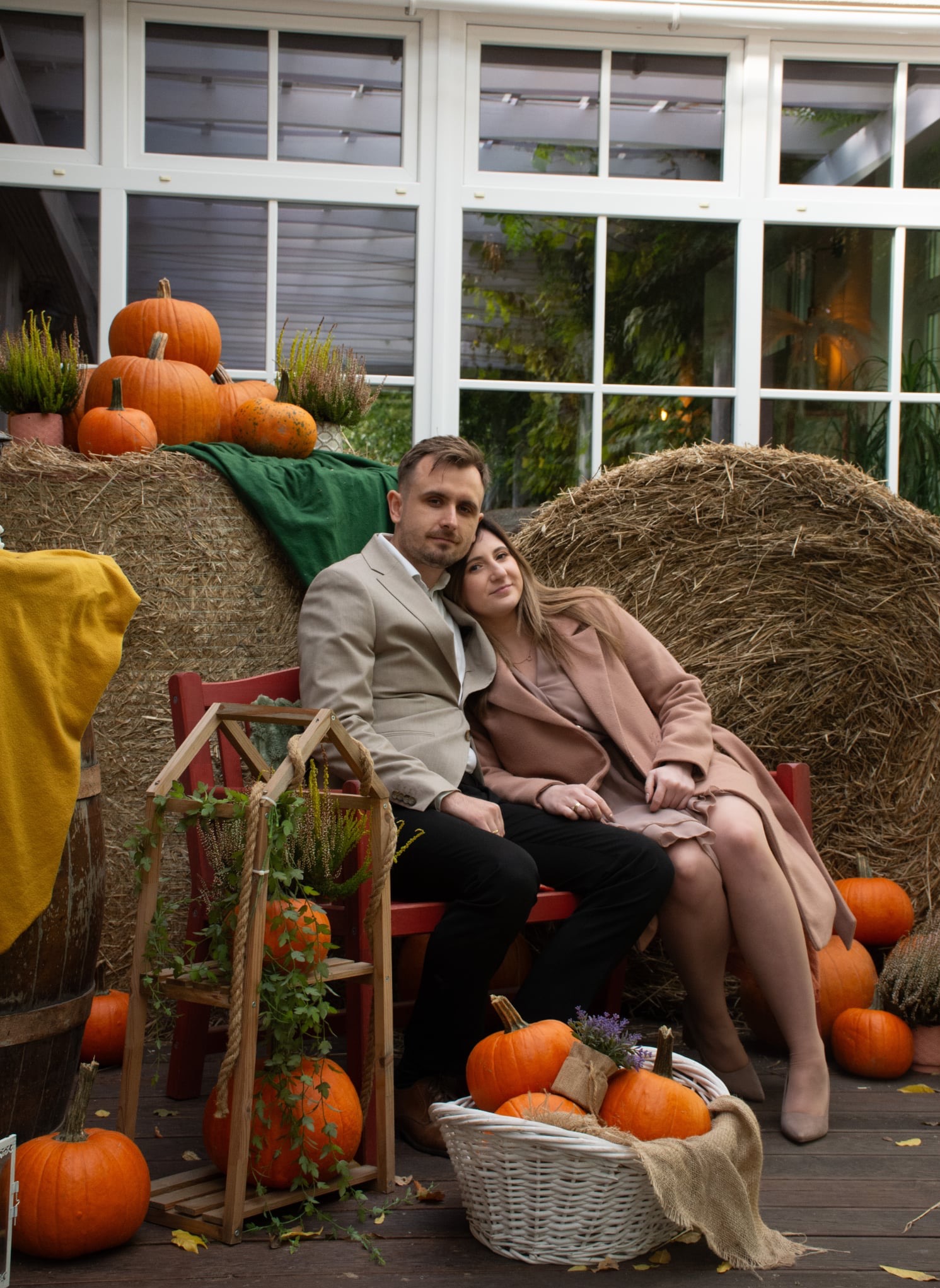Jesienna sesja narzeczeńska: para siedzi na ławce otoczona dyniami i dekoracjami w rustykalnym stylu, na tle siana i okien.
